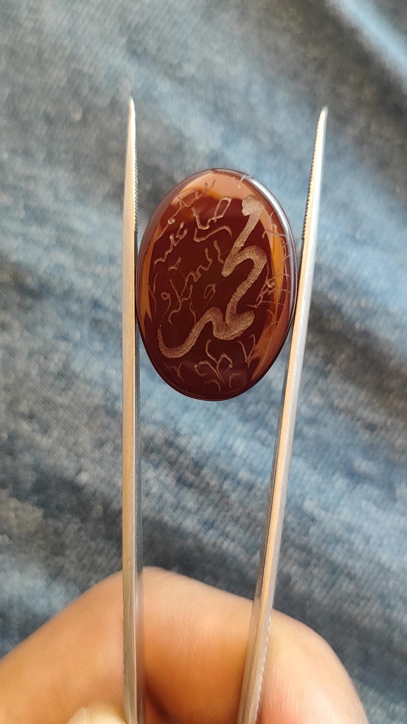 May include: A polished, oval-shaped, reddish-brown gemstone held by tweezers. The stone features an etched, swirling design in a lighter color, possibly depicting calligraphy or a stylized pattern. The background is a blue fabric.