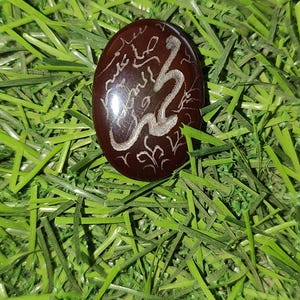 May include: A dark brown, oval-shaped stone with silver-colored calligraphy and a stylized snake design. The stone is set against a backdrop of green artificial grass.