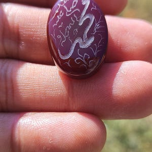 May include: An oval-shaped deep red gemstone featuring intricate white engraved Arabic calligraphy and decorative floral patterns on its smooth, polished surface. The stone has a translucent quality and a rounded, cabochon-style top.