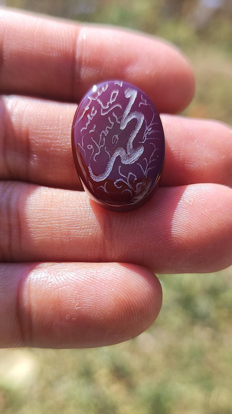 May include: An oval-shaped deep red gemstone featuring intricate white engraved Arabic calligraphy and decorative floral patterns on its smooth, polished surface. The stone has a translucent quality and a rounded, cabochon-style top.