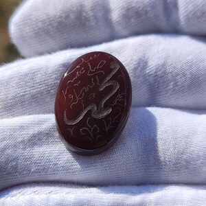 May include: An oval-shaped, deep reddish-brown carnelian gemstone featuring intricate, light-colored engraved Arabic calligraphy on its smooth, polished surface. The stone has a translucent quality and a rounded, cabochon-style top.