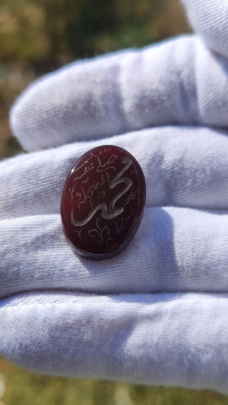 May include: An oval-shaped, deep reddish-brown carnelian gemstone featuring intricate, light-colored engraved Arabic calligraphy on its smooth, polished surface. The stone has a translucent quality and a rounded, cabochon-style top.