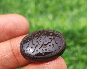 May include: An oval-shaped black gemstone featuring intricate hand-carved Arabic calligraphy and small star-shaped motifs etched into the polished surface. The stone has a smooth, glossy finish and a dark, opaque appearance with detailed engravings.