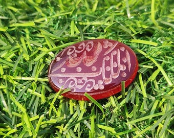 May include: An oval-shaped, deep red gemstone featuring intricate, light-colored engraved Arabic calligraphy across its polished surface. The stone has a smooth, translucent appearance and is displayed as a singular, flat decorative piece.