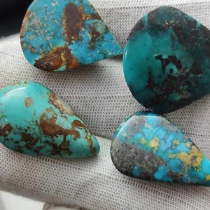 Può includere: Primo piano di cinque pietre preziose di turchese a forma di goccia. Le pietre mostrano un vibrante colore blu-verde con motivi a matrice marrone e nera. Le pietre preziose sono su uno sfondo bianco, evidenziando la loro bellezza naturale.
