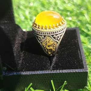 May include: A silver ring featuring a large, oval, yellow gemstone. The ring has a gold-coloured bezel and decorative detailing on the band, including a gold-coloured emblem on the front. The ring is displayed in a black velvet box.