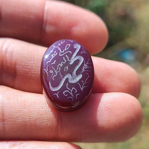 May include: Oval-shaped, deep purple gemstone with white Arabic calligraphy. The stone has a polished surface and is held in a hand. The calligraphy appears to be an inscription or prayer, with decorative floral elements.