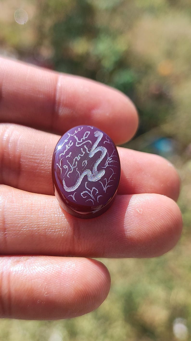 May include: Oval-shaped, deep purple gemstone with white Arabic calligraphy. The stone has a polished surface and is held in a hand. The calligraphy appears to be an inscription or prayer, with decorative floral elements.