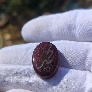 May include: Oval-shaped, burgundy-colored gemstone with silver-colored calligraphy. The stone is held in a white gloved hand. The calligraphy appears to be an inscription or a name. The stone has a polished surface.