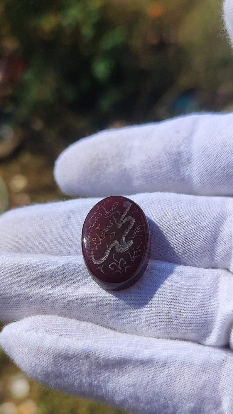 May include: Oval-shaped, burgundy-colored gemstone with silver-colored calligraphy. The stone is held in a white gloved hand. The calligraphy appears to be an inscription or a name. The stone has a polished surface.