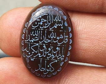 May include: An oval-shaped dark brown gemstone featuring intricate white Arabic calligraphy engraved on its surface. The stone has a smooth, polished finish and is surrounded by a decorative border of small, repeating crescent-shaped indentations.