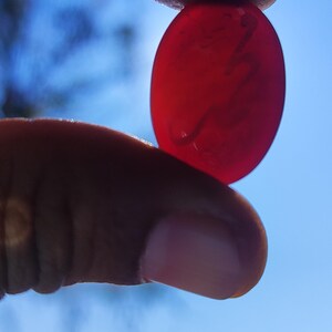 May include: A translucent, oval-shaped, red gemstone is held between two fingers against a bright blue sky. The stone has a smooth surface and appears to be polished. The gemstone's color is a deep, rich red.