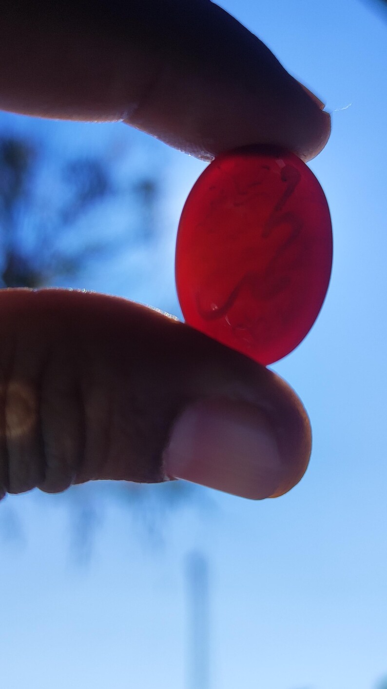May include: A translucent, oval-shaped, red gemstone is held between two fingers against a bright blue sky. The stone has a smooth surface and appears to be polished. The gemstone's color is a deep, rich red.