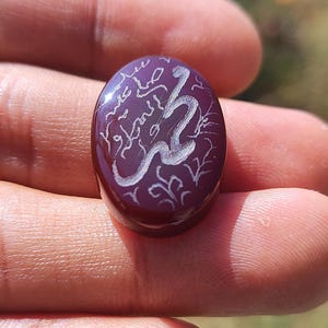 May include: A maroon oval-shaped gemstone with white Arabic calligraphy. The stone is held in a hand, and the text appears to be an inscription. The design includes floral accents. The stone is polished and reflects light.