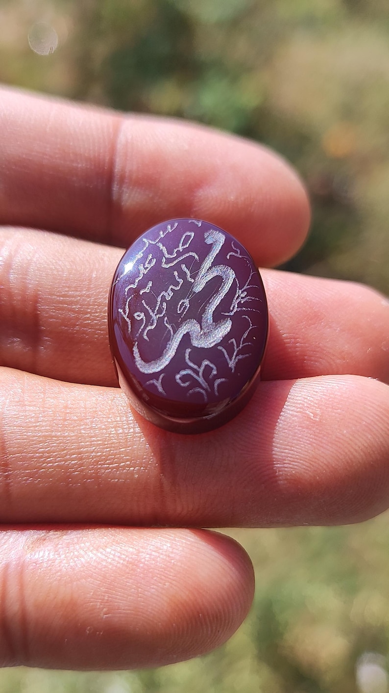 May include: A maroon oval-shaped gemstone with white Arabic calligraphy. The stone is held in a hand, and the text appears to be an inscription. The design includes floral accents. The stone is polished and reflects light.