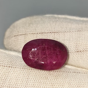May include: A faceted, oval-shaped ruby gemstone. The gem is a deep, saturated red colour with internal inclusions. The ruby is set against a white fabric background, highlighting its colour and clarity. The gemstone is a focal point.