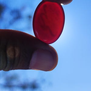 May include: A translucent, oval-shaped, red gemstone is held between two fingers against a bright blue sky. The stone is smooth and polished, catching the sunlight. The image is a close-up, highlighting the gem's color and form.