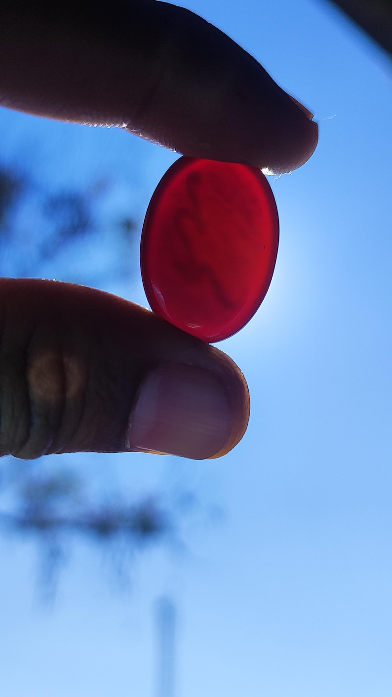 May include: A translucent, oval-shaped, red gemstone is held between two fingers against a bright blue sky. The stone is smooth and polished, catching the sunlight. The image is a close-up, highlighting the gem's color and form.