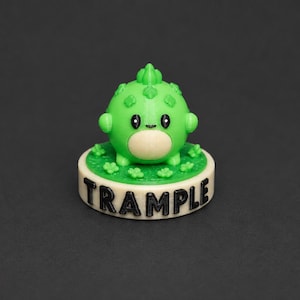 May include: A small, round, green creature figurine with a light-colored belly and black eyes, standing on a circular base. The base is green with small leaf shapes and features the word TRAMPLE in bold, black, raised capital letters on the side.