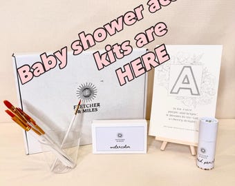 May include: A selection of baby shower activity kits. The image shows a white box, watercolour paints, paintbrushes, a small easel with a floral design, and a pencil. Text on the box reads "Fletcher & Miles". The text "Baby shower activity kits are HERE" is also visible.