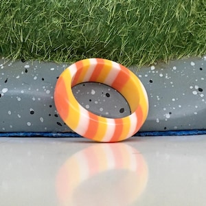 Puede incluir: Un anillo de silicona de rayas naranja, blanco y amarillo brillante. El anillo está sobre una superficie gris con motas blancas y negras.