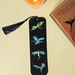 Puede incluir: Un marcapáginas negro con un acabado brillante y cuatro siluetas de dragones holográficos iridiscentes. El marcapáginas tiene un borla negra.