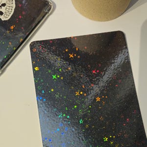 Op de afbeelding: Een zwarte kaart met een holografische afwerking met een patroon van regenboogsterren op een zwarte achtergrond.