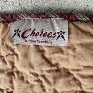 May include: Close-up of a quilted fabric with a label that reads "Choices In Hand Creations." The fabric is tan with dark blue stitching and a plaid border in red, green, and gold.