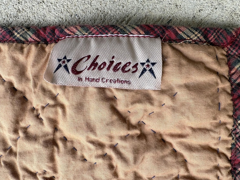 May include: Close-up of a quilted fabric with a label that reads "Choices In Hand Creations." The fabric is tan with dark blue stitching and a plaid border in red, green, and gold.