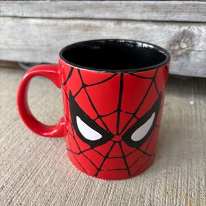 May include: A red ceramic mug featuring a black spiderweb design and white eye shapes, reminiscent of the Spider-Man character. The mug has a black interior and a red handle, suitable for hot or cold beverages.