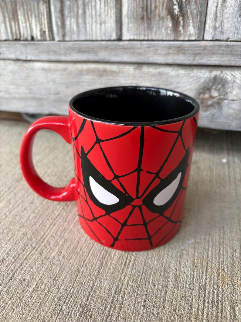 May include: A red ceramic mug featuring a black spiderweb design and white eye shapes, reminiscent of the Spider-Man character. The mug has a black interior and a red handle, suitable for hot or cold beverages.