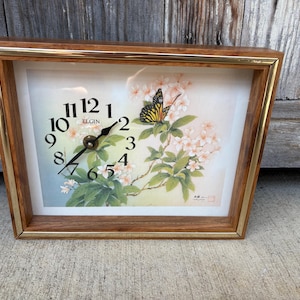 May include: A rectangular wall clock with a wood-grain frame and gold-tone trim. The clock face features black Arabic numerals, the Elgin brand name, and a printed illustration of a monarch butterfly resting on a branch of light pink flowers.
