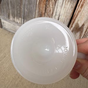Może przedstawiać: Okrągła podstawa kubka z białego szkła mlecznego z wytłoczonym napisem Heat Proof. Sp&oacute;d kubka jest gładki i błyszczący, wykazuje p&oacute;łprzezroczystość typową dla szkła mlecznego, z literami odciśniętymi bezpośrednio w szklanej powierzchni przedmiotu.