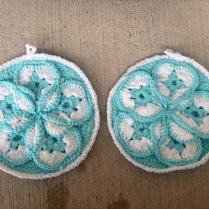 May include: Two round, crocheted potholders. Each potholder features a floral design in white and turquoise, with a white border and a small loop for hanging. The potholders are handmade.