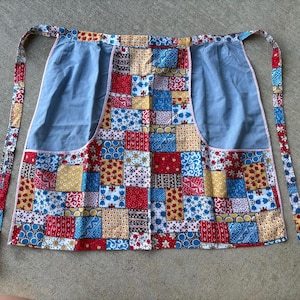 May include: A half apron featuring a patchwork design of various red, blue, and yellow floral and geometric patterns. The apron includes two large, solid light blue pockets on the front and long fabric ties on each side for securing the garment.