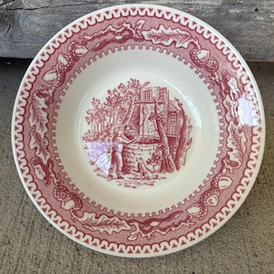 May include: A round, off-white ceramic bowl with a red border featuring a repeating pattern of acorns and leaves. The center of the bowl has a red illustration of a well scene with figures and a dog.