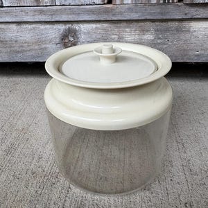 May include: A clear glass storage canister with a wide, off-white plastic lid. The lid features a raised circular handle in the center and a wide, flat rim that extends beyond the glass base. The container has a cylindrical shape and smooth walls.