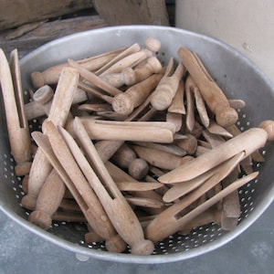 May include: A collection of wooden clothespins in a silver metal colander. The clothespins are light brown and have a classic design with a spring-loaded clamp. The colander is round with small holes and is filled with many clothespins.