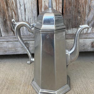 May include: A silver-toned metal coffee pot with a hexagonal body, a curved handle, and a matching lid with a pointed finial. The vessel features a long, slender spout and a small circular hallmark stamped on the upper front panel of the body.
