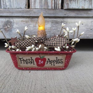 May include: A red metal bread pan with white speckles, decorated with a painted apple and the text "Fresh 5¢ Apples". The pan is filled with white faux baby's breath and a lit candle.