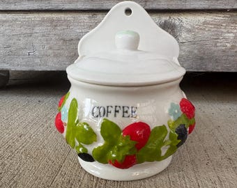 May include: White ceramic coffee jar with a lid and a decorative hanging back. The jar is adorned with red strawberries, green leaves, and the word "COFFEE" in black lettering. The jar is a charming kitchen accessory.