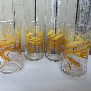 May include: Set of four clear drinking glasses with a gold wheat stalk design. The glasses are tall and cylindrical, with a slightly flared top. The wheat design is repeated on each glass.