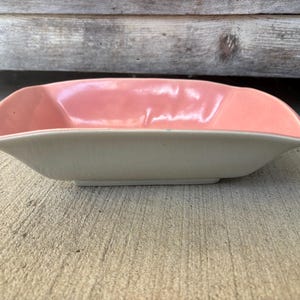 May include: A vintage, rectangular serving dish with a light pink interior and a cream-colored exterior. The dish has gently curved sides and a slightly raised base, showcasing a mid-century modern aesthetic. The dish is likely made of ceramic or porcelain.