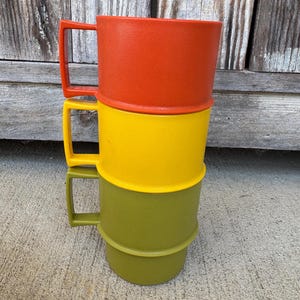 May include: A stack of three plastic mugs featuring a textured finish. The top mug is bright orange, the middle mug is vibrant yellow, and the bottom mug is olive green. Each mug has a square-shaped handle and a slightly tapered cylindrical body.