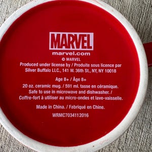 May include: A red ceramic mug with the Marvel logo and text. The mug is 20 oz. and safe for microwave and dishwasher use. The text includes the Marvel website and copyright information.
