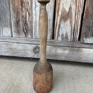 May include: A tall, cylindrical wooden kitchen tool with a rounded handle top and a wider, heavy base. The wood features a natural, weathered light brown finish with visible grain patterns, signs of wear, and a smooth, tapered neck section.