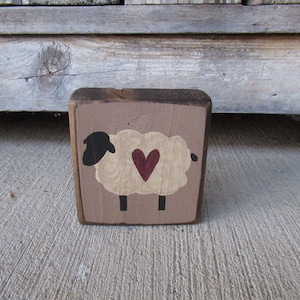 May include: A wooden block with a hand-painted sheep design. The sheep has a cream-colored body, a black head, and a red heart. The block is painted with a light brown background and has a rustic, handmade appearance.