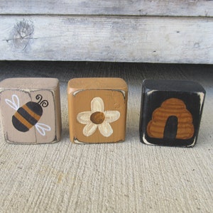 May include: Three wooden blocks with painted designs. The first block is beige with a black and yellow bee. The second block is brown with a white flower. The third block is black with a brown beehive.