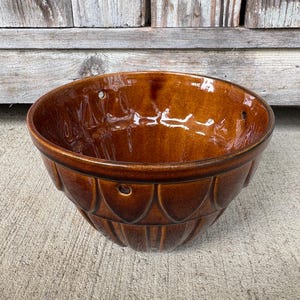 May include: A round, glazed ceramic bowl in a deep amber-brown color. The exterior features a repeating vertical petal or scalloped pattern. Three small, evenly spaced circular holes are positioned near the upper rim for hanging purposes.