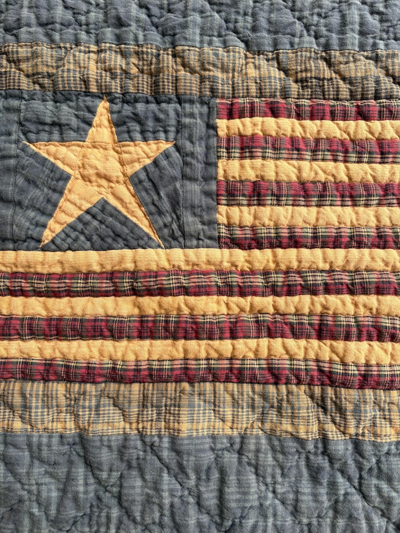 May include: A quilted textile featuring a design reminiscent of the American flag. The quilt showcases a blue field with a gold star and alternating stripes of gold and red plaid. The fabric has a textured, quilted appearance.